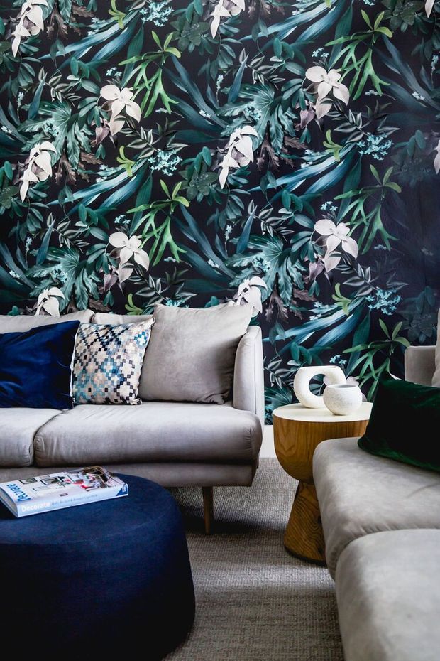 A richly styled living room featuring bold dark botanical wallpaper with tropical leaves and pale flowers, a light grey sofa and armchair, patterned and dark velvet cushions, a rounded navy ottoman, a sculptural wooden side table, and neutral textured carpeting. The room has a layered, dramatic and luxurious feel with strong botanical patterning and contrasting jewel-toned accents.