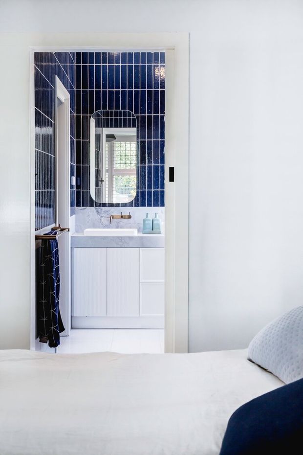 A bright, refined bathroom viewed from a bedroom through an open doorway. The bathroom features deep navy vertically arranged wall tiles with white grout, a rounded-rectangle mirror, pale marble-look benchtop, white fluted vanity cabinetry, a white basin, and brushed brass tapware. The foreground shows part of the white bed and navy bedding, clearly establishing the direct bedroom-to-bathroom connection.