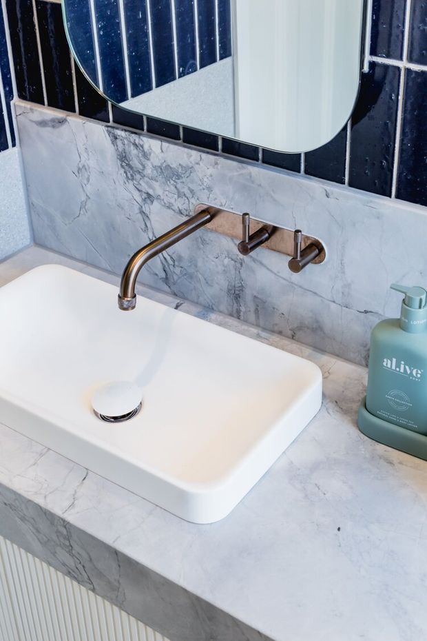 Close-up of a refined bathroom vanity featuring a white vessel basin set into a pale grey marble-look countertop and backsplash. A brushed bronze wall-mounted tap contrasts with glossy dark navy rectangular wall tiles, while a rounded-corner mirror and green soap dispenser add finishing details.