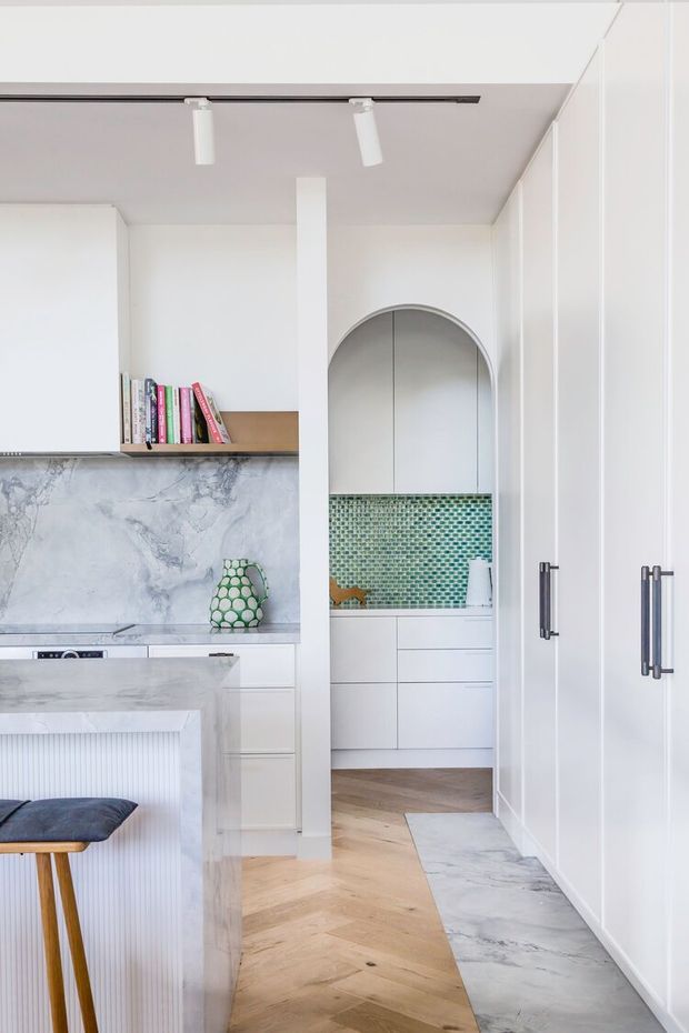 Bright, refined contemporary kitchen with white cabinetry, a pale veined stone splashback and island, herringbone timber flooring, and a compact green mosaic-tile wet-area or pantry visible through an arched opening. Integrated tall storage lines the right wall, with minimalist track lighting and a partially visible bar stool.