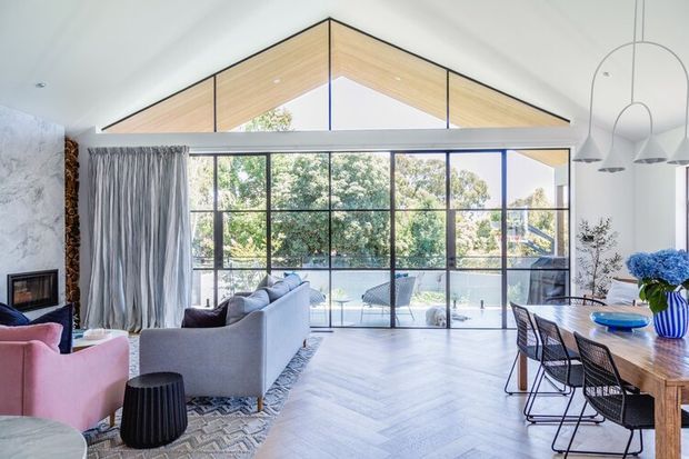 Bright open-plan living and dining space with a dramatic pitched ceiling and expansive black-framed glazing opening to an outdoor area. The room includes pale herringbone timber flooring, a large grey sofa, pink accent armchairs, a timber dining table with woven black chairs, sculptural pendant lighting, and a stone-clad fireplace wall.