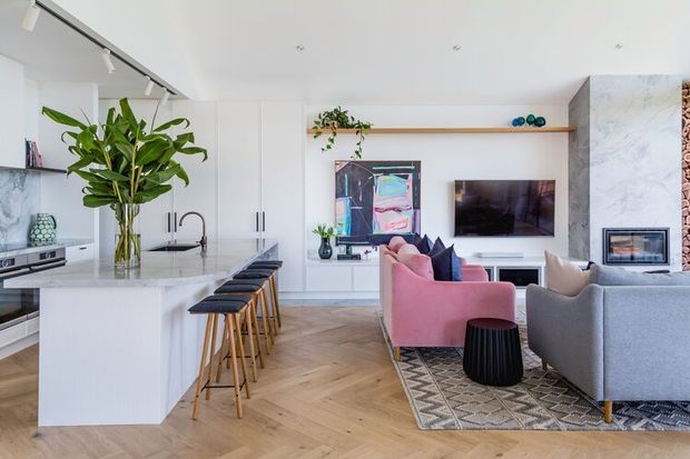 Bright open-plan contemporary living area with a white kitchen island and integrated seating, timber herringbone flooring, built-in white cabinetry, television, fireplace, and a mix of grey and blush-pink sofas. Marble-look surfaces, indoor greenery, track lighting, and a patterned rug add contrast and warmth.