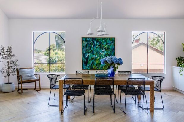 Bright contemporary dining room with a long timber dining table surrounded by black woven chairs, sculptural white pendant lights, pale herringbone flooring, white walls, and large arched-grid windows. Indoor plants and a colourful artwork add natural and blue-green accents.