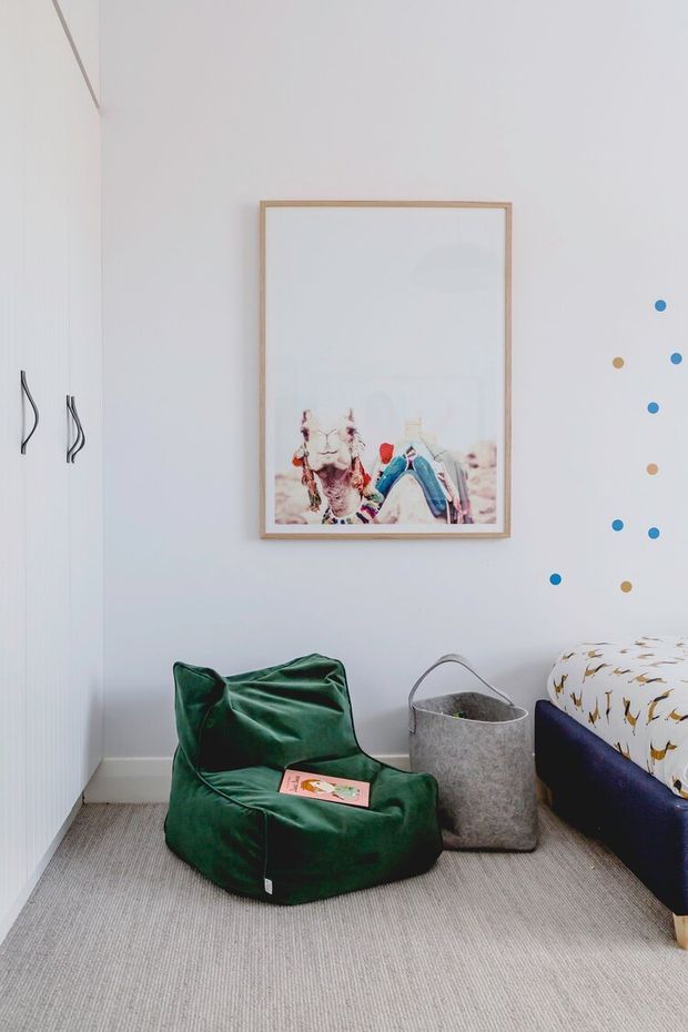 A bright, compact bedroom corner with white walls and built-in white cabinetry, a grey carpeted floor, and a low navy upholstered bed partially visible at right. A dark green beanbag chair, grey wastebasket, framed artwork, and colourful circular wall decals create a playful yet minimal children’s room setting.