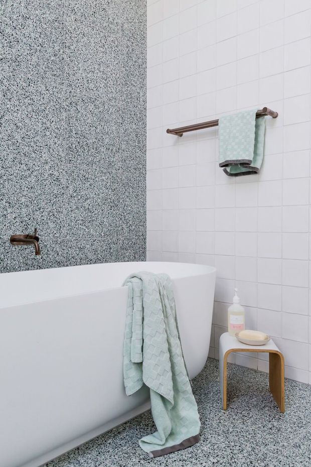 A bright, contemporary bathroom featuring a freestanding white bathtub, white square tiled walls, and a terrazzo-style speckled wall and floor finish. Copper-toned tapware and towel rails complement a pale mint-green towel, with a small timber stool holding soap and bath accessories.