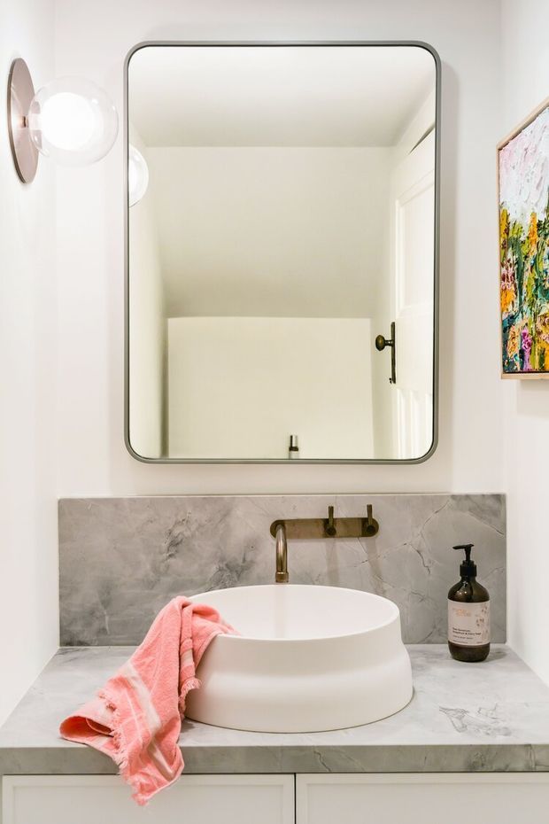 A refined bathroom vanity featuring a white vessel basin set on a grey marble benchtop, with a matching marble splashback, brushed brass wall-mounted tapware, and a rounded rectangular mirror. A spherical wall light and colourful artwork add decorative accents to the otherwise light, neutral space.