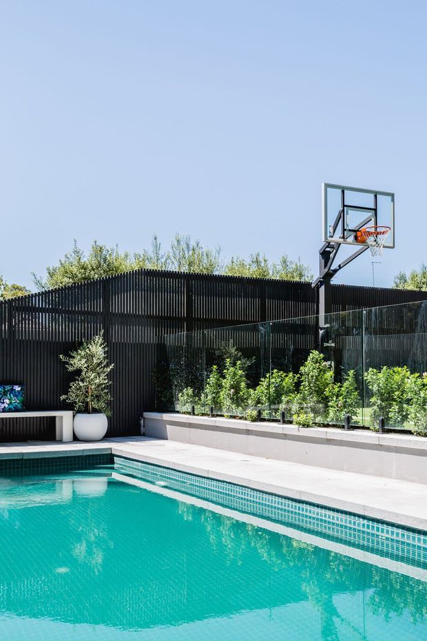 Bright outdoor residential pool area with turquoise tiled water, pale concrete paving, a raised planter filled with greenery, a black vertical-slat privacy fence, and a glass basketball backboard and hoop. The scene has a contemporary, landscaped backyard character under a clear blue sky.