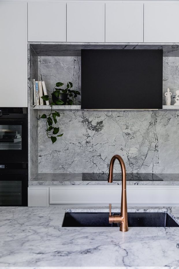 A close-up view of a contemporary kitchen featuring extensive grey-veined marble surfaces, white flat-panel cabinetry, a matte black rangehood and integrated black ovens. A copper-toned gooseneck tap projects over a dark undermount sink in the marble island, while a small trailing plant and books add detail to the splashback niche.