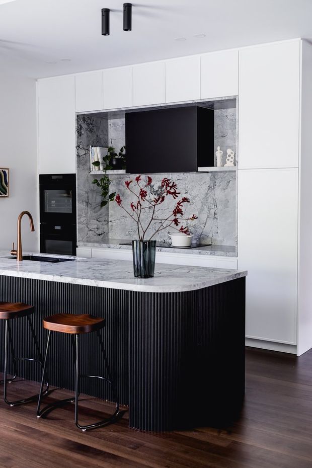 A refined contemporary kitchen with a large marble island featuring a black vertically fluted base, integrated sink and bronze-toned curved tap. White full-height cabinetry surrounds a dark television or display niche framed in veined marble, with integrated black appliances. Two timber-seated black bar stools sit at the island, and dark timber flooring adds warmth. The palette is predominantly white, black, grey marble and dark brown timber, with restrained decorative greenery and red branches.