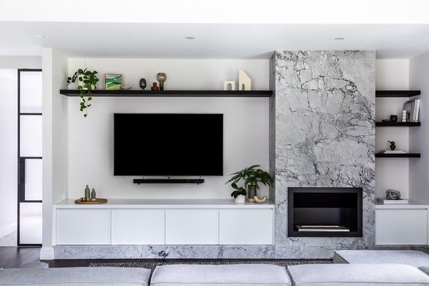 Bright contemporary living room featuring a full-width white built-in media wall with low cabinetry, open display shelving, a wall-mounted television, and a dramatic grey-white veined stone fireplace surround. Minimal recessed lighting, dark floating shelves, indoor plants, and a pale sectional sofa create a clean, refined interior.