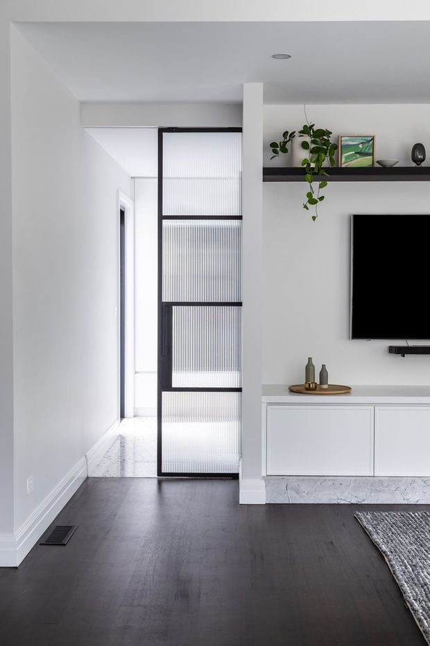 A bright, minimalist living room with white walls and ceiling, dark timber flooring, and a white built-in media console. A wall-mounted television sits beneath a long black display shelf with plants and simple decor. A black-framed ribbed translucent glass sliding door separates the room from a hallway, while a narrow vertical column and recessed ceiling light add architectural structure.