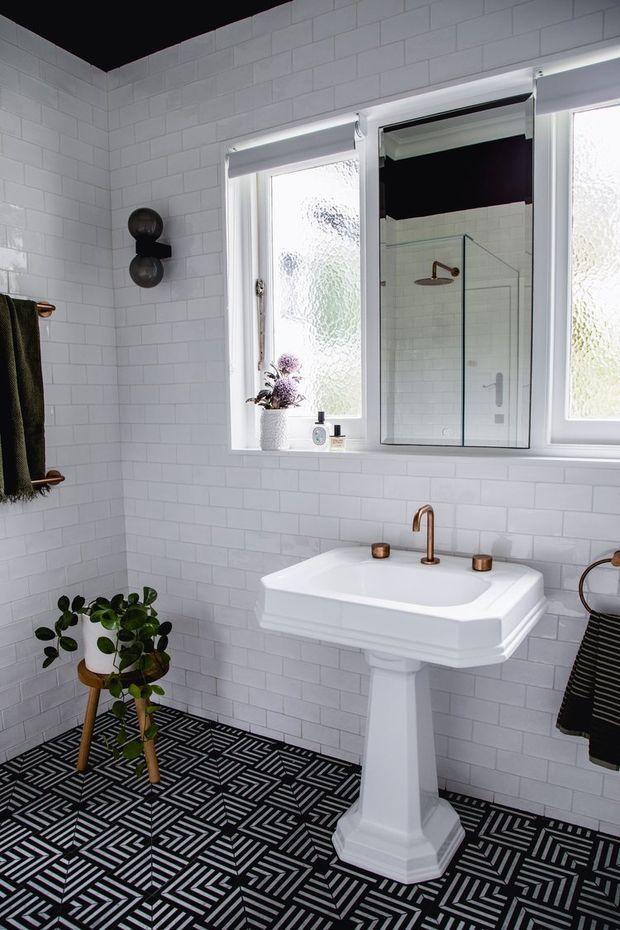 A bright bathroom with white subway-tiled walls, a black-and-white geometric patterned tile floor, and a traditional white pedestal basin with copper-toned tapware. Frosted windows and a mirrored cabinet reflect a glass shower enclosure. Small plants, towels, and dark decorative accents add contrast.