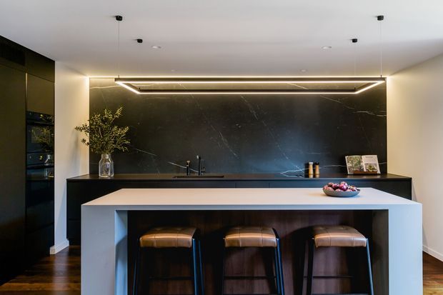 A sophisticated contemporary kitchen featuring a large island with integrated seating for three, dark timber cabinetry, a dramatic black veined stone splashback, and a linear pendant-style LED feature light. Built-in appliances are concealed within tall dark cabinetry, while warm timber flooring and leather stools add contrast.