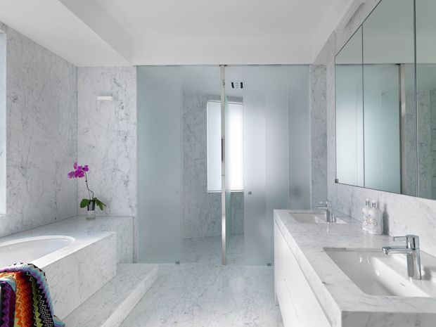 Bright, luxurious contemporary bathroom clad extensively in white-grey veined marble, featuring a freestanding bathtub at left, a walk-in glass shower at center, and a double vanity with mirrored wall cabinets at right. The space has clean white surfaces, chrome fixtures, and a colourful towel with orchid providing small accents.
