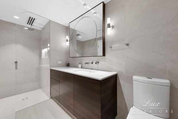 Contemporary bathroom with a frameless glass walk-in shower, large pale stone-look wall and floor tiles, a floating dark timber vanity with integrated white basin, round wall mirror, modern wall sconces, and a toilet. The space is clean, refined, and neutral with strong contrast from the dark joinery.