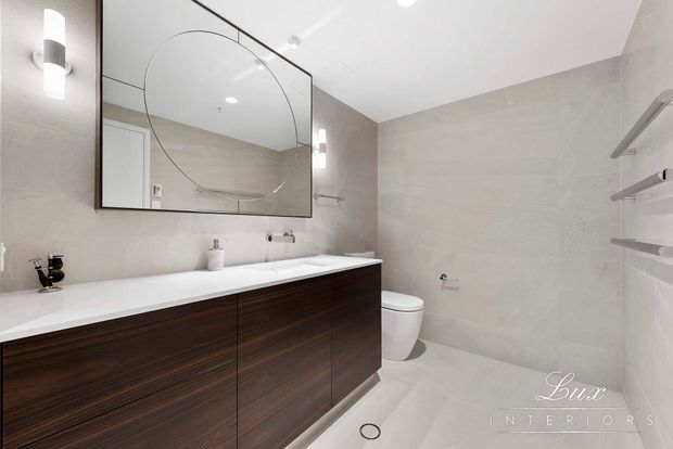 Contemporary bathroom with a long floating dark timber vanity, white benchtop and basin, wall-mounted toilet, large frameless curved-edge mirror, pale stone-look wall and floor finishes, recessed shelving, and integrated wall lighting.