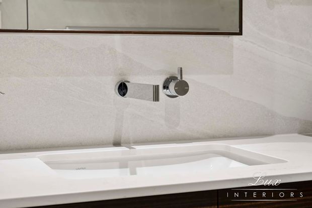 Close-up of a contemporary bathroom vanity featuring a white integrated basin and benchtop, wall-mounted chrome tapware, pale stone wall finish, and dark timber cabinetry below. A framed mirror is partially visible above.