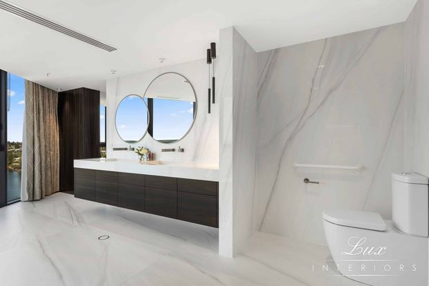 Contemporary bathroom with a long dark timber floating vanity, white benchtop, twin round mirrors, wall-mounted black cylindrical sconces, and a separate toilet area. Large-format white marble-look wall and floor surfaces create a bright, polished finish, with full-height glazing and curtains visible at the side.