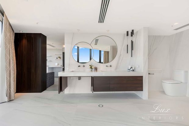 Luxury contemporary bathroom with a large floating dark timber double vanity, white stone benchtop, twin round mirrors, wall-mounted black tapware and sconces, and pale marble-look floor and wall surfaces. A freestanding-style toilet is visible in a partially enclosed zone, with a dark timber joinery wall and bedroom or dressing area beyond.