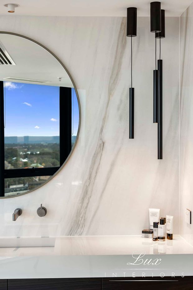 Contemporary bathroom vanity with a large round mirror, pale marble-look wall finish, white integrated basin, dark minimalist pendant lights, and black cabinetry. A coastal city view is reflected in the mirror.