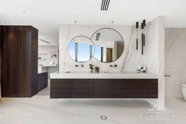 Contemporary bathroom vanity with a long dark timber floating cabinet, white stone benchtop, large round mirrors, marble-look wall surfaces, integrated lighting and a partial view of a toilet. A bedroom or dressing area is visible through the opening on the left, but the bathroom is the primary space.