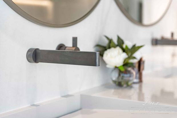 Close-up of a contemporary bathroom vanity with a wall-mounted dark metal faucet, white tiled wall, pale countertop, round mirrors, and a small floral arrangement. The scene has a clean, refined, softly styled appearance.