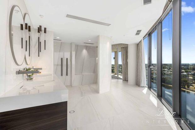 Bright, luxurious contemporary bathroom with a floating dark-wood vanity, white stone surfaces, mirrored wall, sculptural pendant lighting, and a spacious open shower area. Floor-to-ceiling glazing and sheer curtains provide expansive city views and abundant natural light.