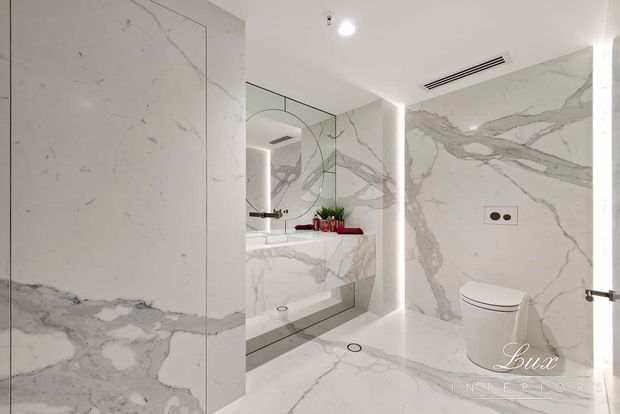 A bright, luxurious contemporary bathroom clad almost entirely in dramatic white marble with bold grey veining. The space features a wall-hung toilet, floating marble vanity with integrated basin, large mirrored cabinet, frameless glass shower partition, concealed linear air vent, and recessed strip lighting.