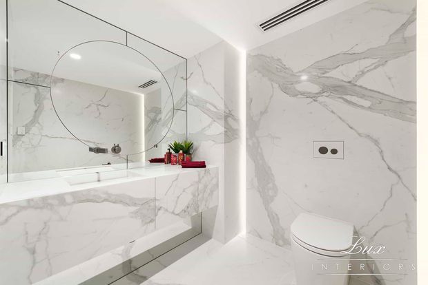 A bright, luxurious contemporary bathroom clad extensively in white marble-look surfaces with grey veining. The room features a freestanding or partially visible bathtub behind a clear glass screen, a large mirrored wall with a curved etched detail, integrated LED lighting, a wall-hung toilet, and minimal chrome fixtures. A small red decorative arrangement provides contrast on the bath ledge.