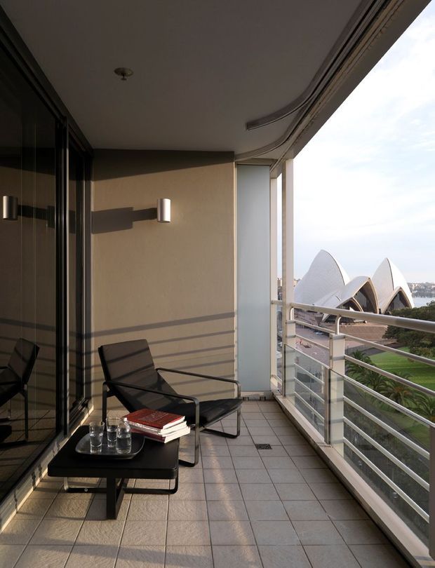 A covered apartment balcony or terrace with a tiled floor, glass sliding doors, metal-and-glass balustrade, wall-mounted lighting, and modern outdoor lounge furniture. The Sydney Opera House is clearly visible across the water through the open side, with landscaped grounds below.