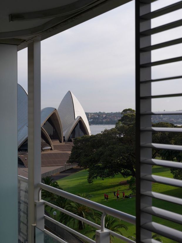 View from a building through horizontal louvres and a balustraded opening, overlooking the distinctive sail-like roof shells of the Sydney Opera House, landscaped lawns, harbour water and the opposite shoreline. The frame is dominated by architectural glazing, structural columns and external shading elements, creating a strong framed urban landmark vista.