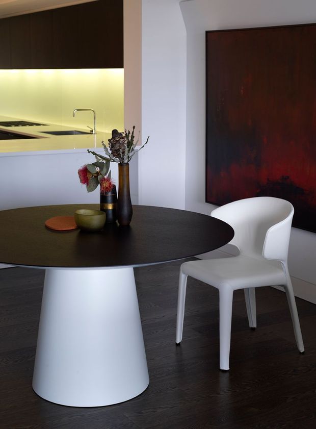 A refined contemporary dining area with a large round dark dining table on a sculptural white pedestal base, paired with a modern white armchair. The space features dark timber flooring, a partially visible kitchen with dark cabinetry and a pale illuminated splashback, white walls, and a large dark red and black abstract artwork. Decorative vases and dried botanical stems provide a restrained accent on the table.
