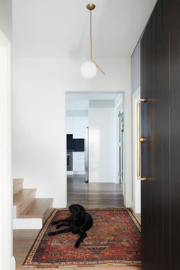 Bright, narrow residential entry hall with white vertical wall panelling, pale timber stairs, dark timber entry door with a long brass handle, and a contemporary brass-and-glass pendant light. A richly patterned red Persian-style rug anchors the floor, with a black dog resting on it. The hallway opens toward a white kitchen and adjoining circulation spaces in the background.