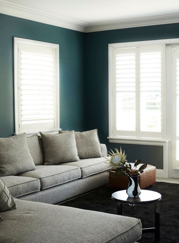 A refined living room with deep blue-green painted walls, white plantation shutters and detailed white crown moulding. Two large grey upholstered sofas frame a small round black coffee table, with a dark rug and understated botanical arrangement creating a calm, polished atmosphere.