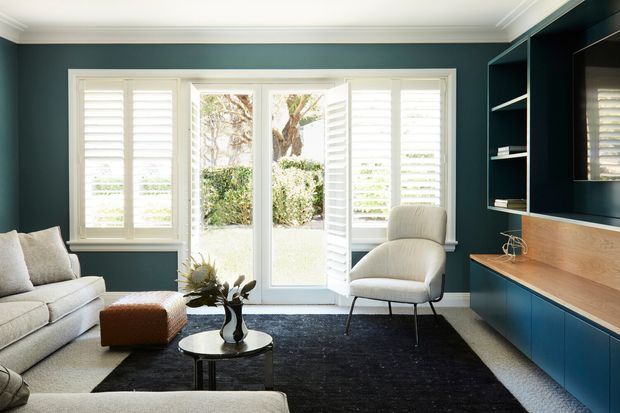 A refined living room with deep teal walls, white plantation shutters and French doors opening to a landscaped garden. The room includes a pale upholstered sofa, sculptural lounge chair, small ottoman, round coffee table, black rug, and a built-in dark blue media wall with open shelving and warm timber cabinetry.