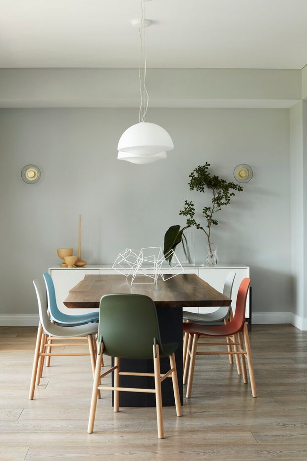 A refined dining room with a pale grey-green wall, light timber flooring, and a large dark timber dining table surrounded by colourful mid-century-style chairs. A white sideboard, sculptural white pendant light, small wall lights, ceramics, and indoor plants complete the calm, contemporary setting.
