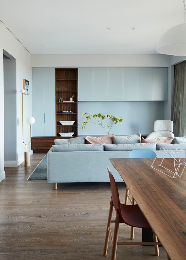 A bright, refined open-plan living and dining area with pale blue-grey built-in cabinetry, integrated shelving, a large light blue sectional sofa, timber dining table and chairs, and warm timber flooring. Minimal recessed ceiling lights, a sculptural pendant, soft curtains, and restrained decor create a clean contemporary atmosphere.