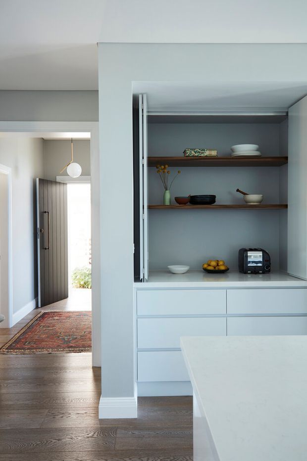 A refined, light-filled kitchen with pale blue-grey built-in cabinetry, open dark timber shelving, white benchtops, and integrated lower drawers. A compact appliance and decorative crockery sit on the shelves. The foreground includes the edge of a large kitchen island, while an open doorway leads to an entry hall with timber flooring and a patterned runner.