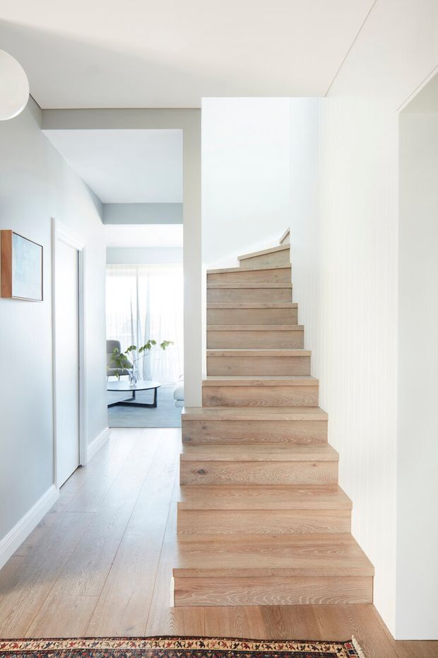 Bright, minimalist residential hallway with a prominent straight-run timber staircase rising through the centre-right of the frame. Pale oak-toned stair treads and matching light timber flooring contrast with smooth white and very light grey walls. A small living area with a coffee table and plants is visible at the end of the corridor, while a patterned rug adds a subtle accent in the foreground.