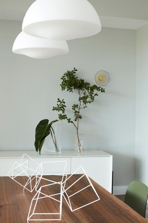 A bright, minimalist dining area with a walnut-toned timber dining table, white geometric wire-frame chairs, and a pale green upholstered chair. A white built-in sideboard sits against a soft light-grey wall, decorated with clear glass vases and leafy branches. Large white dome pendant lights hang overhead, and a small circular wall light provides an additional focal point.