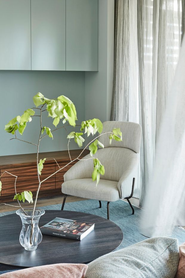 A softly lit, contemporary living room featuring a sculptural upholstered armchair beside a round dark wood coffee table. Pale blue-green built-in cabinetry and wall panelling line the background, with sheer full-height curtains over a large window. A leafy branch in a glass vase, a design book, layered rugs, and neutral cushions create a calm, refined interior.