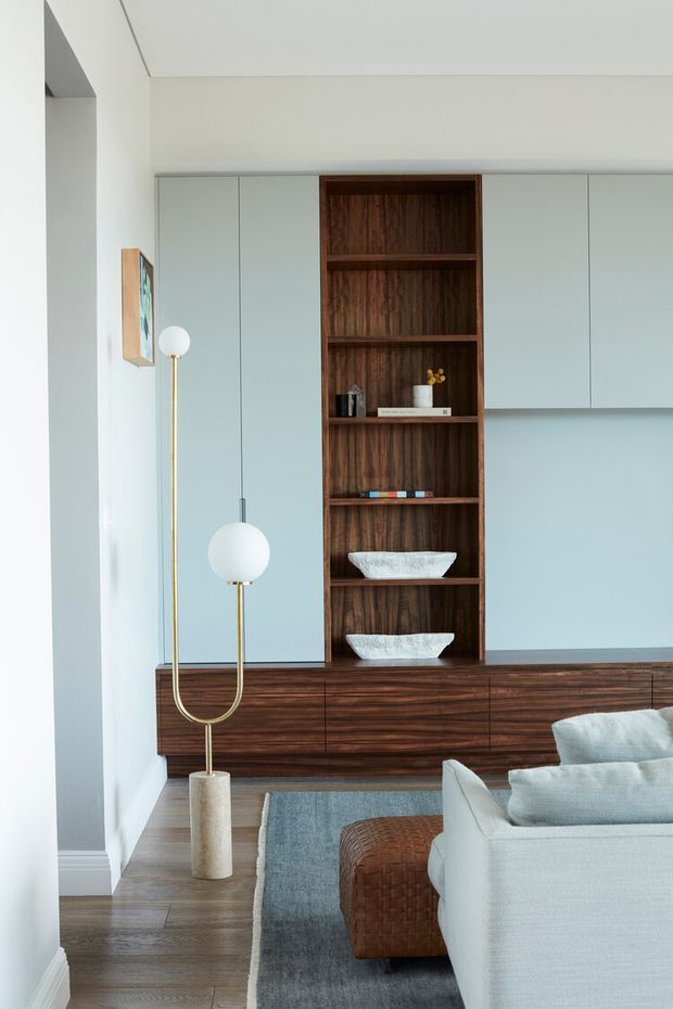 A refined living room with a pale blue-grey built-in media and storage wall, featuring a rich dark timber base cabinet and central open shelving. A sculptural brass floor lamp, textured white accessories, pale upholstered seating, a muted blue-grey rug, and timber flooring create a calm, contemporary interior.