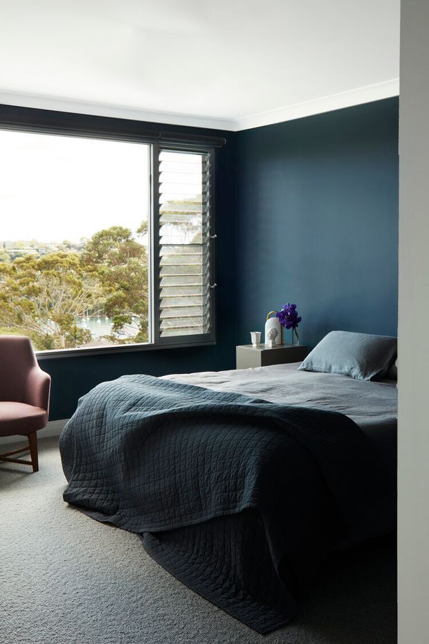 A contemporary bedroom with deep navy-blue walls, white ceiling and trim, a large louvred window overlooking trees and water, and a neatly made bed dressed in dark blue-grey quilted bedding. A small bedside table with flowers and a mauve upholstered chair complete the calm, moody interior.