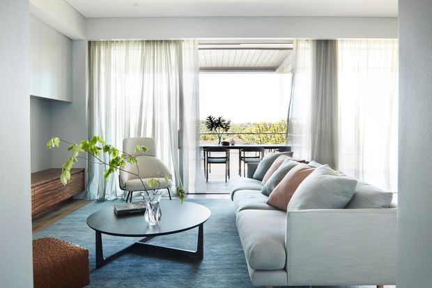 Bright contemporary living room with a large pale sectional sofa, round black coffee table, soft blue rug, sheer full-height curtains, built-in wall cabinetry, and timber storage. Glazing opens onto a balcony or terrace with outdoor dining and a leafy outlook.