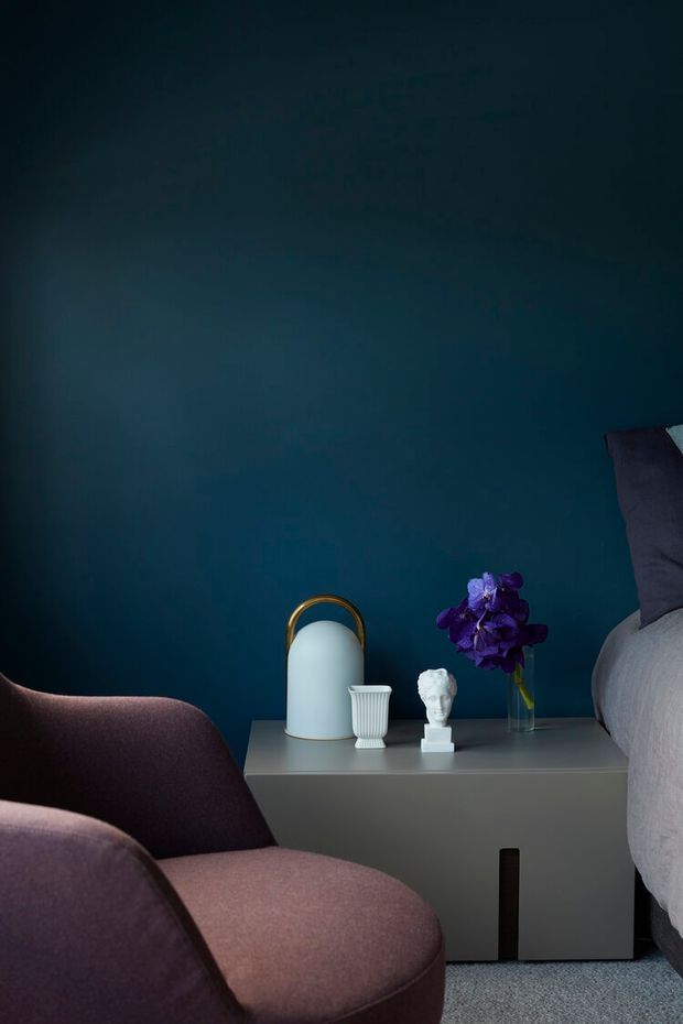 A moody bedroom vignette featuring a deep teal painted wall, a grey bedside table with a sculptural white lamp, pleated vase, classical bust, and purple flowers. A mauve upholstered armchair occupies the foreground beside the edge of a bed with neutral bedding. The room has soft, low lighting and a refined, contemporary decorative style.