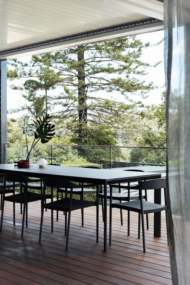 A contemporary indoor-outdoor dining area opening onto a timber deck, with a long dark dining table and matching chairs positioned beside full-height glazing. A glass balustrade frames a leafy treetop outlook, while a covered ceiling and sheer curtain provide shelter and privacy. The space has a restrained black, timber, and green palette with abundant natural light.