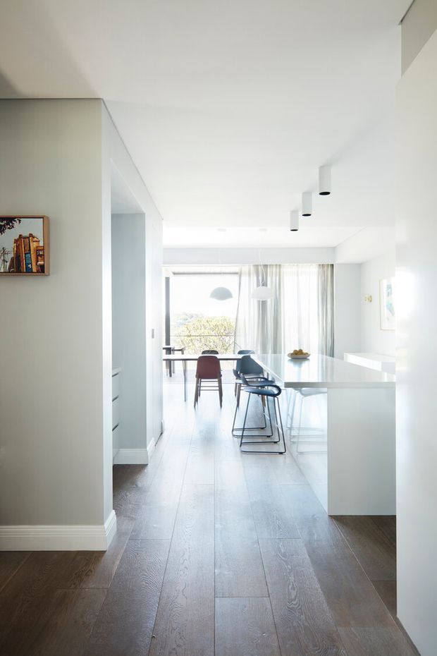 Bright open-plan interior viewed from a hallway or entry area, looking toward a contemporary kitchen and dining space. The room features white walls and cabinetry, dark timber flooring, a pale island bench, a dining table with mixed chairs, sheer curtains, pendant lights, and abundant natural light from large glazing.