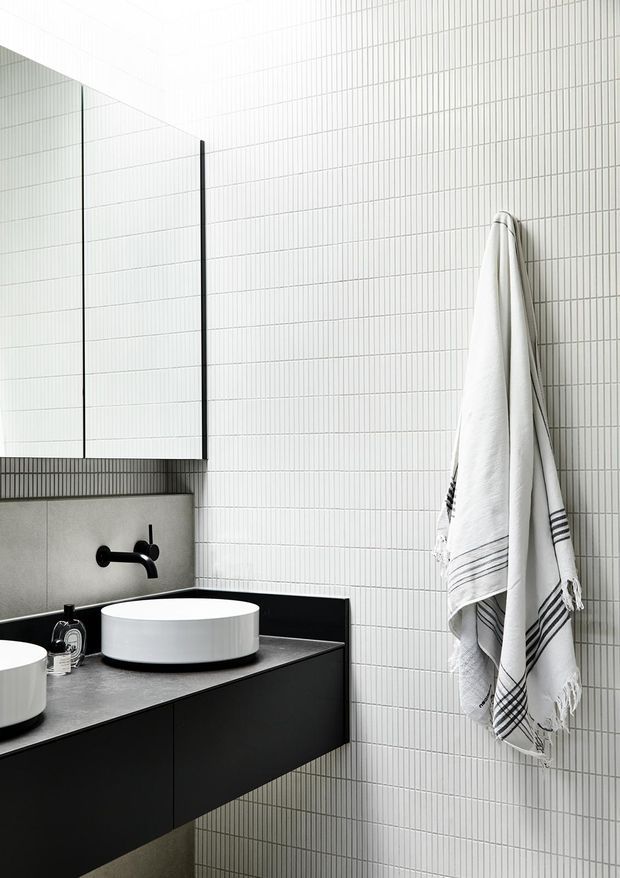 Contemporary bathroom vanity with two round white vessel basins set on a dark floating vanity. The walls are clad in small white grid-pattern tiles, complemented by matte black wall-mounted taps, mirror detailing, and black cabinetry. A white striped towel hangs on the tiled wall.