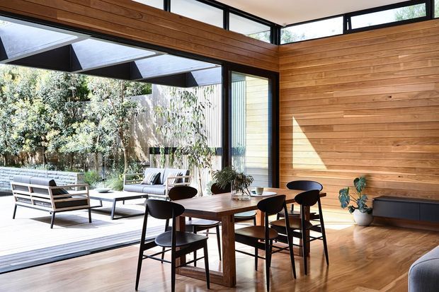 Bright contemporary open-plan dining and living area with warm horizontal timber wall cladding and timber flooring. A large glazed opening connects the interior to a landscaped outdoor terrace, with a timber dining table and chairs in the foreground and lounge seating beyond. High-level clerestory glazing and exposed dark window frames bring in abundant natural light.