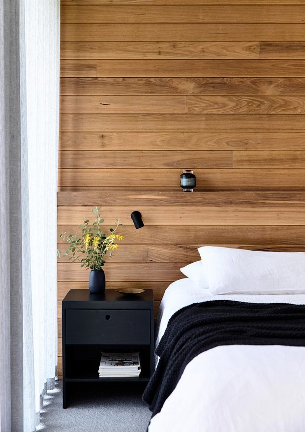 A refined bedroom vignette featuring a full-height horizontal timber feature wall behind the bed, with warm natural wood grain and integrated black wall lighting. A black bedside table with open shelving, a dark vase of yellow flowers, white bedding, and a textured black throw create a high-contrast, contemporary look. Grey carpet and sheer curtains appear at the side.